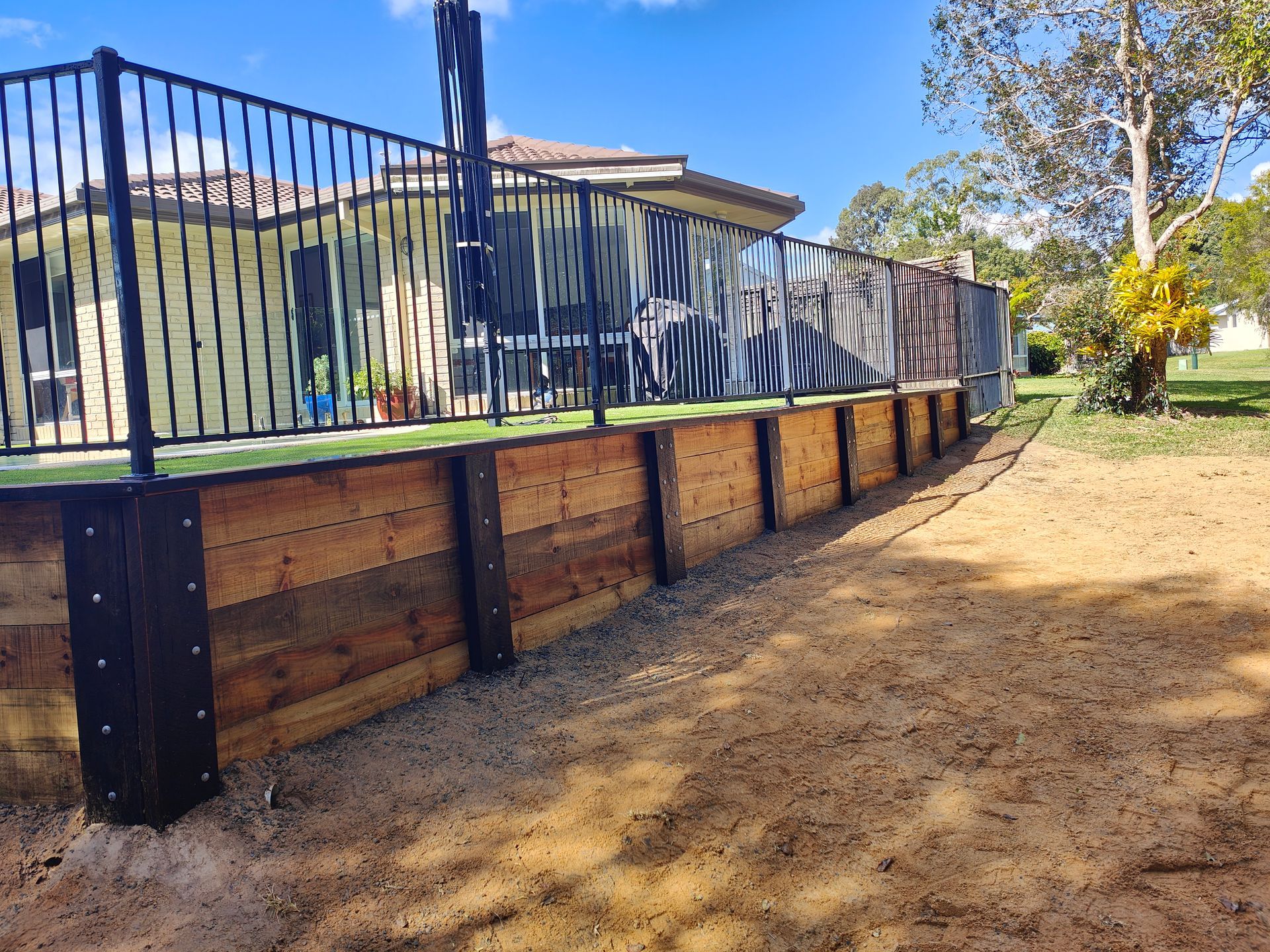 Raised Wooden Retaining Wall With Black Metal Fence Along A Sandy Yard Under A Blue Sky — Quality Fences 4 U in Gympie, QLD