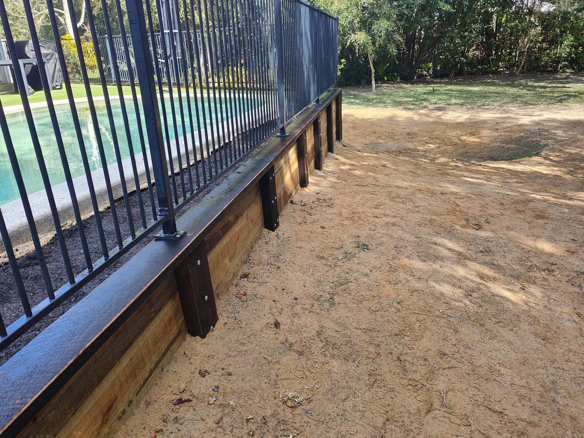 Black metal fence with wooden base along the edge of a pool, set in a sandy yard — Quality Fences 4 U in Gympie, QLD