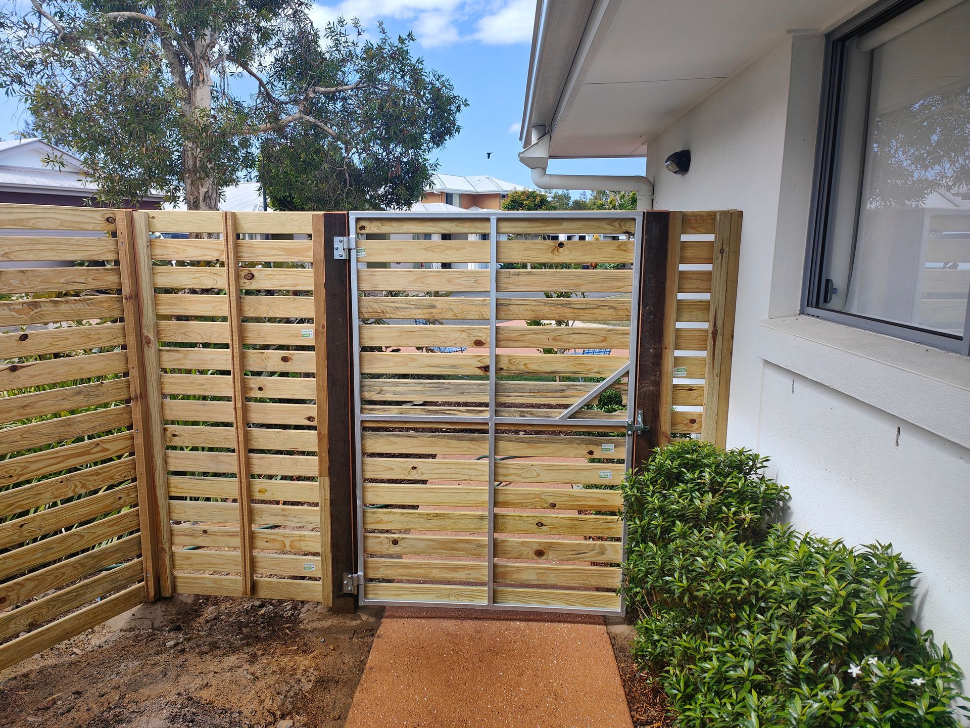 Wooden Slat Gate Beside A White House, With A Small Shrub And Tree In A Gravel Yard — Quality Fences 4 U in Gympie, QLD