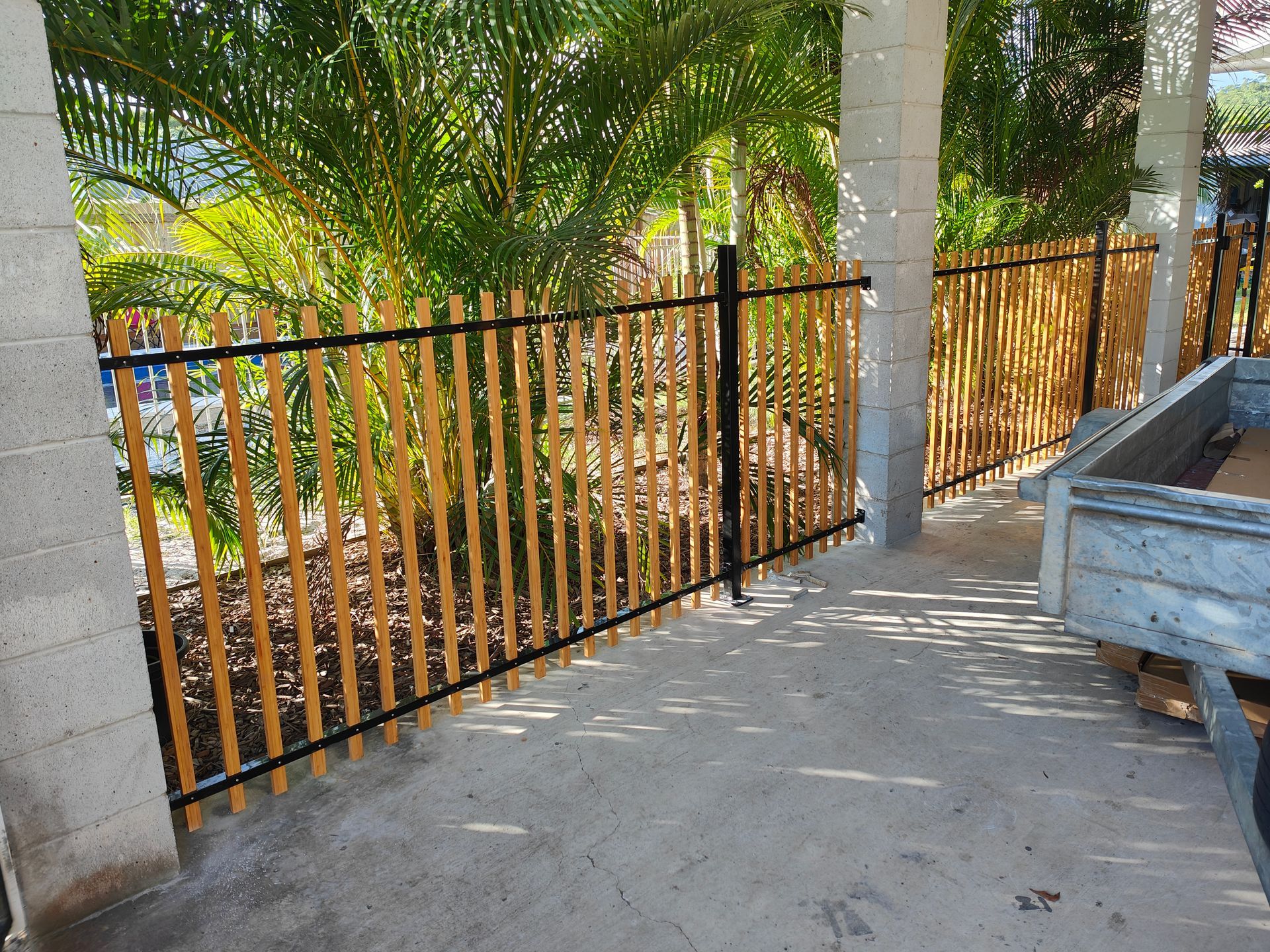 Black Metal Fence Beside A Concrete Walkway Under Palm Trees, With A Gray Utility Cart On The Right — Quality Fences 4 U in Gympie, QLD