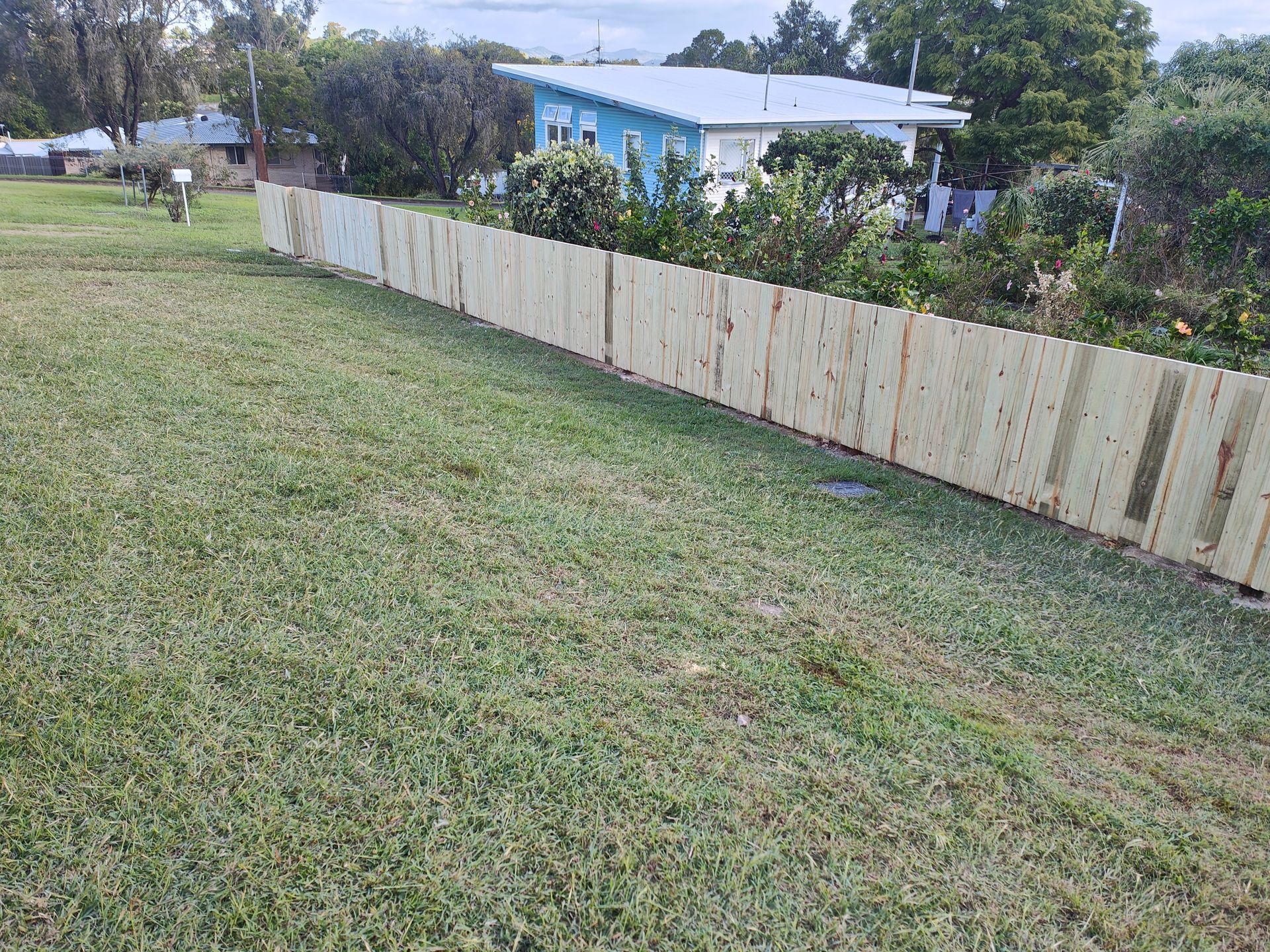 Grassy Yard Beside A Weathered White Fence, With A Blue House And Trees In The Background — Quality Fences 4 U in Gympie, QLD