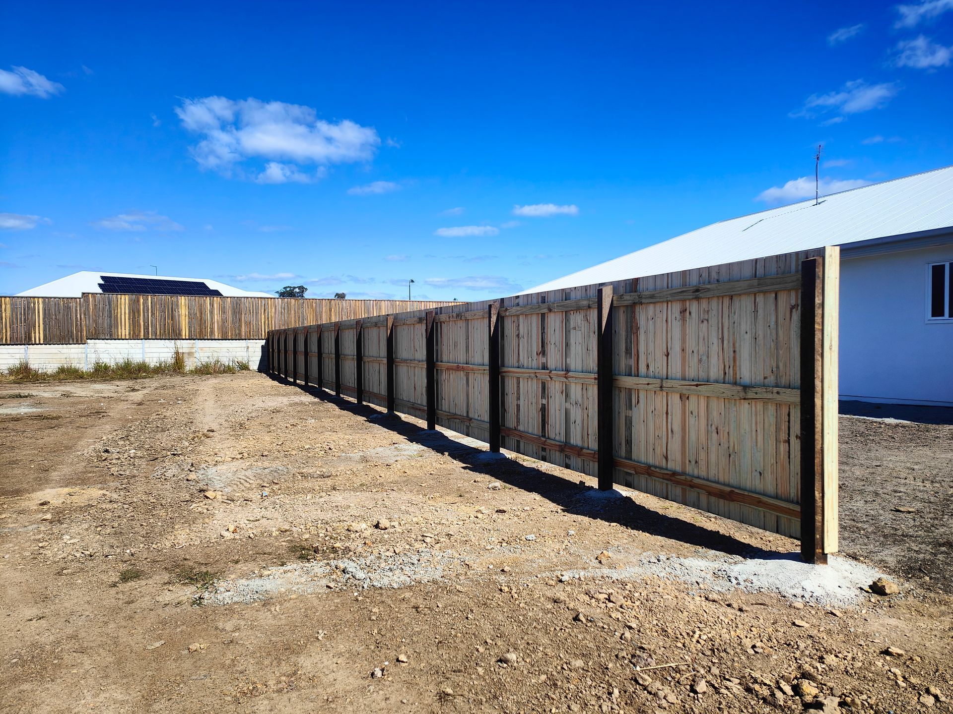 Long Wooden Fence Along A Dirt Lot Beside A White Building Under A Blue Sky — Quality Fences 4 U in Gympie, QLD