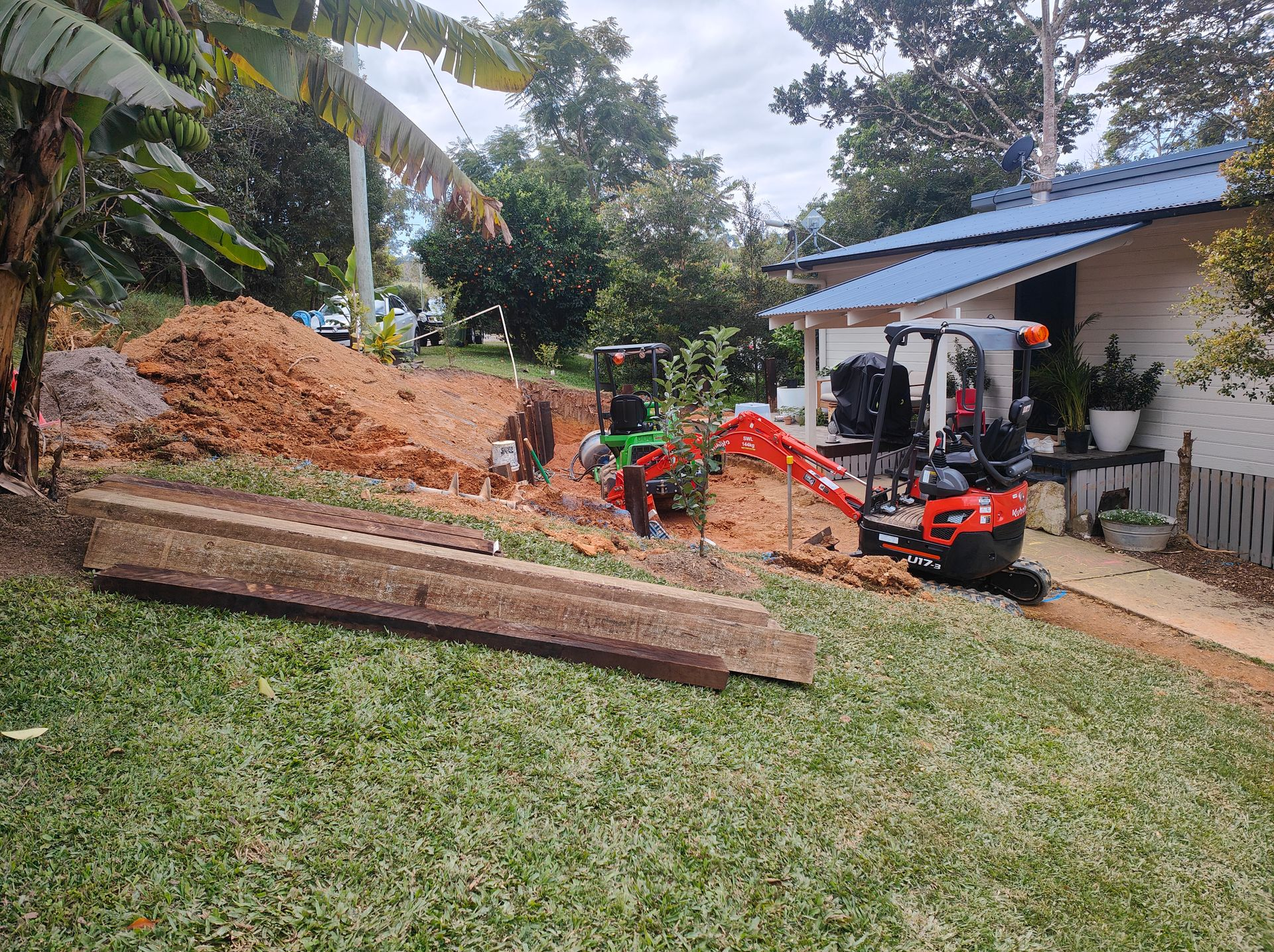 Small Excavator And Workers Moving Dirt Beside A House In A Yard With Palm Trees And Stacked Lumber — Quality Fences 4 U in Gympie, QLD