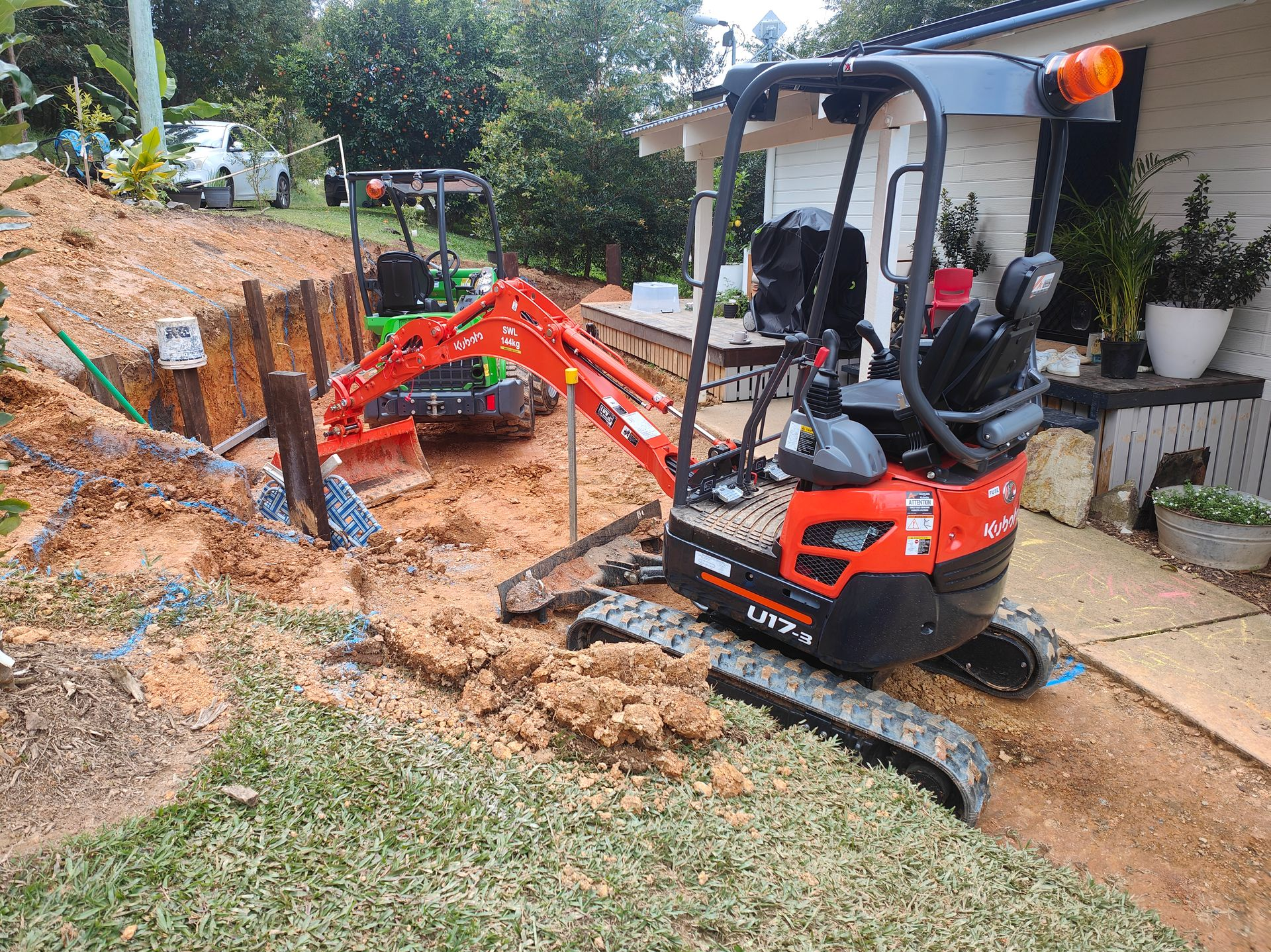 Small Red Excavator Digging Soil Beside A House On A Sloped Yard, With A Worker Nearby — Quality Fences 4 U in Gympie, QLD