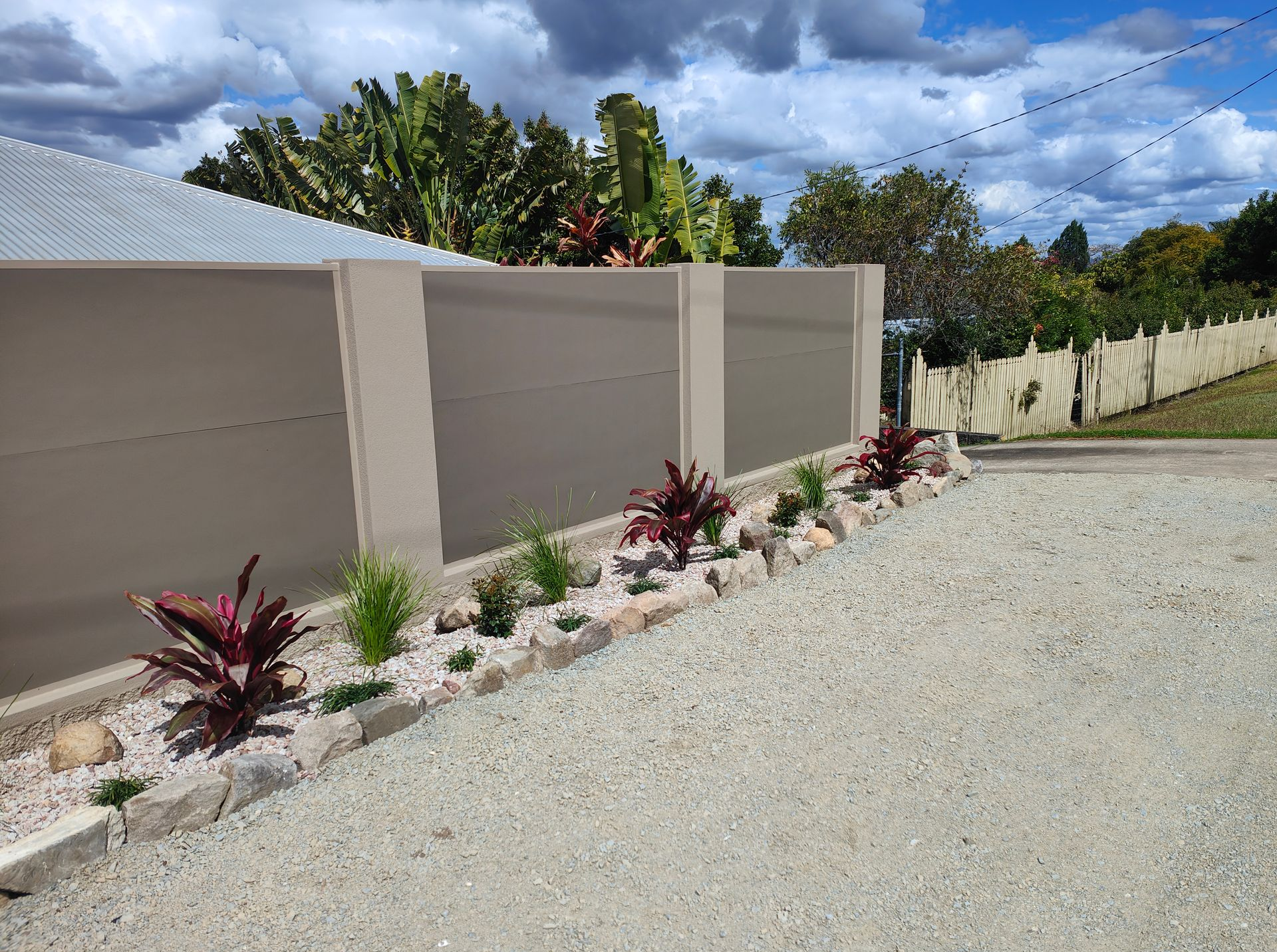 Gravel Driveway Beside A Beige Fence And Landscaped Flower Bed Under A Cloudy Sky — Quality Fences 4 U in Gympie, QLD