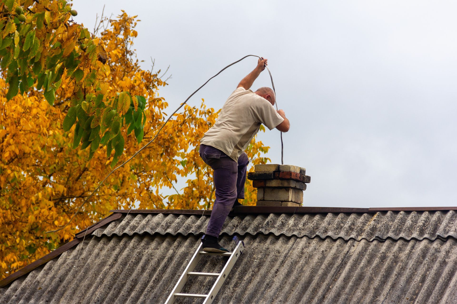 A man is standing on a ladder on top of a roof cleaning a chimney.