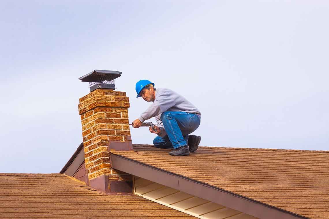 Contractor builder with blue hardhat on the roof caulking chimney – Macungie, PA - Anytime Todd's Chimney Sweeping
