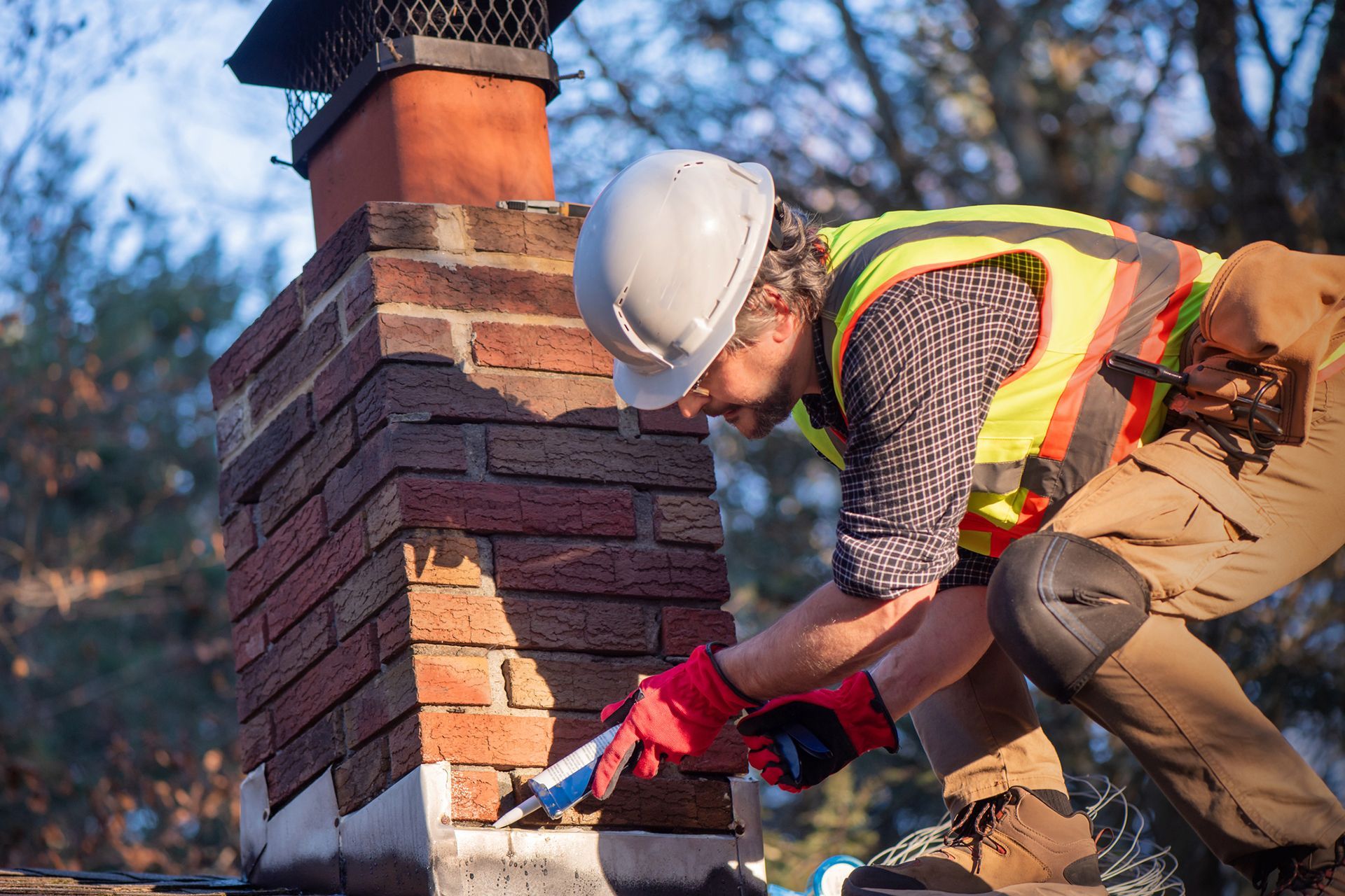 A construction worker in safety gear applies sealant to a brick chimney on a rooftop.