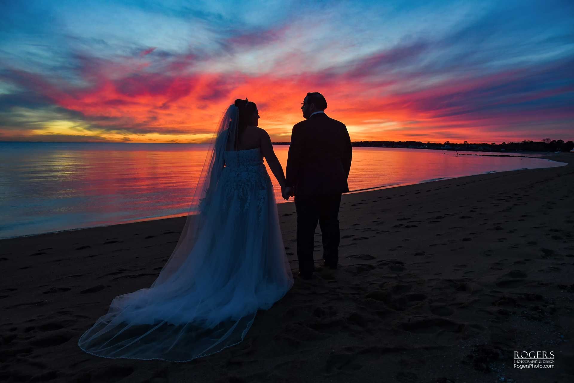 Silhouetted couple standing on a beach at sunset beside a vivid orange and purple sky
