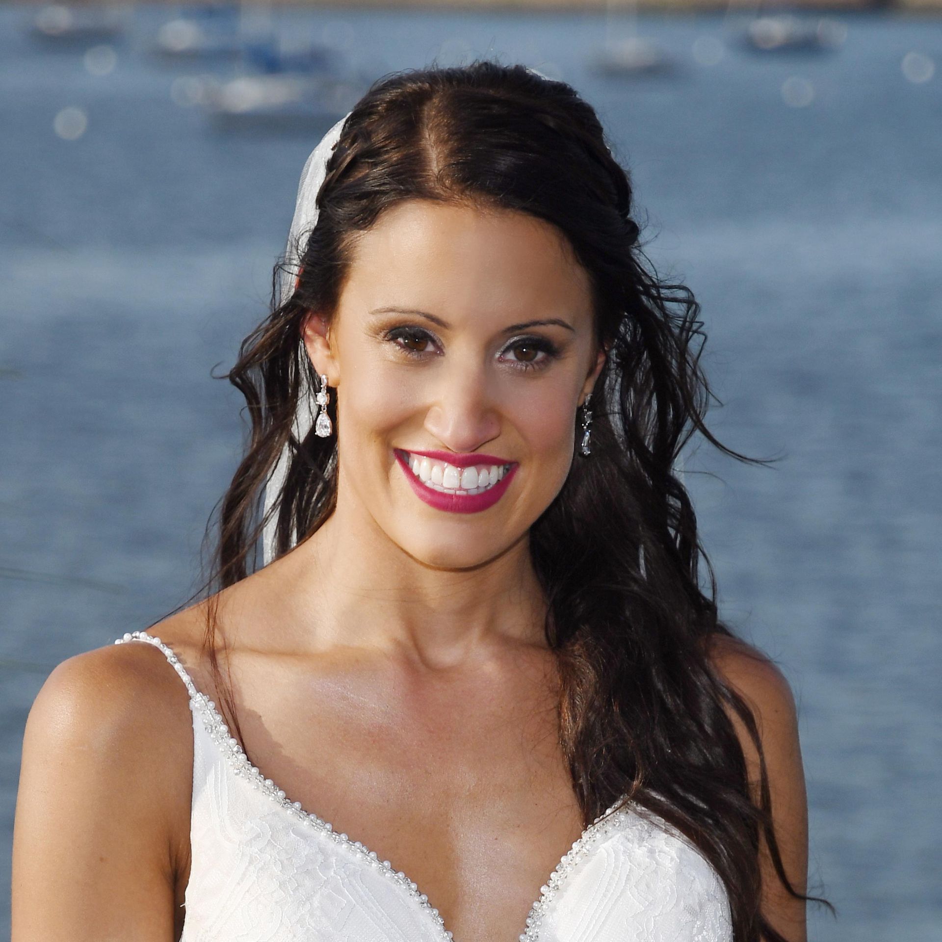 Smiling Bride in a white dress standing by the water, with dark hair and bright pink lipstick.
