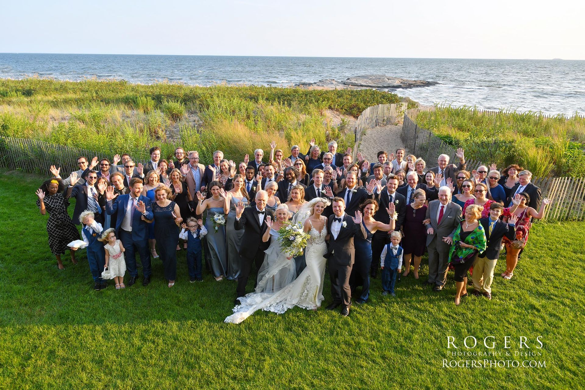 A large group of people at a wedding reception posing for a picture with the ocean in the background at Madison Beach Hotel wedding photographed by Rogers Photography in Connecticut. 