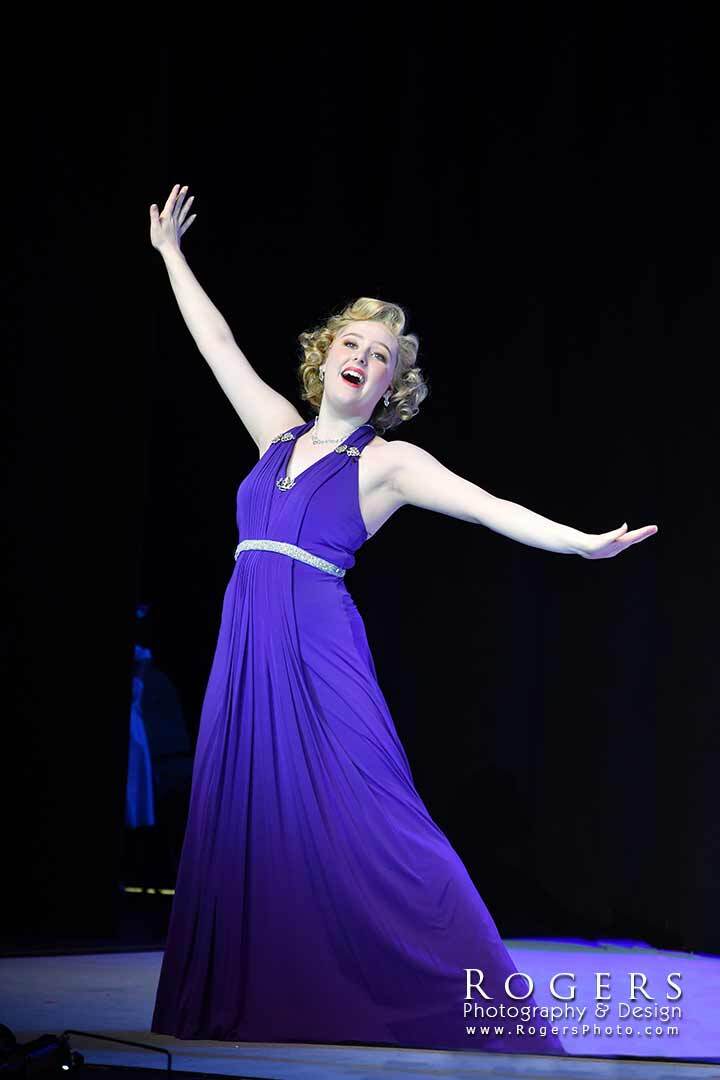 Woman in a purple dress dancing on stage with arms raised against a dark background for the production of Anything Goes.