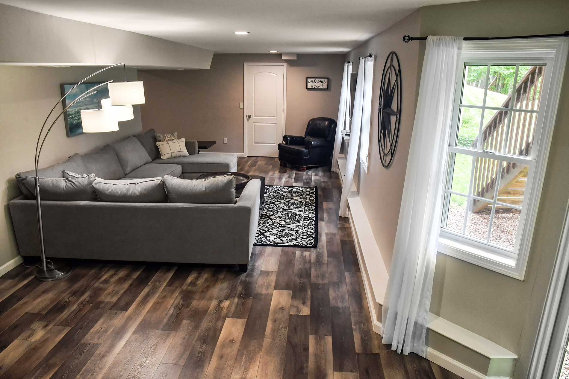 Modern living room with gray sectional sofa, dark wood floor, patterned rug, and large window with sheer curtains
