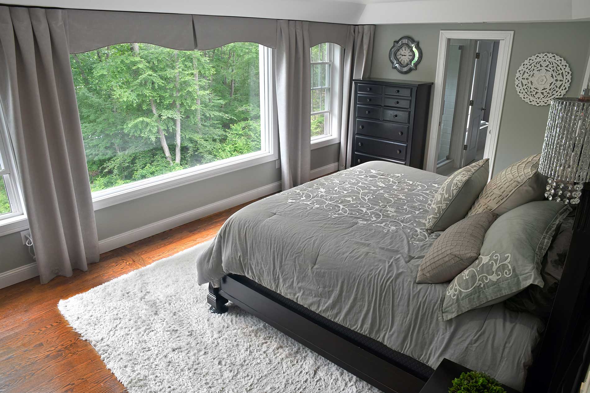 Bright bedroom with large window, gray bed, white rug, and dresser beside greenery outside