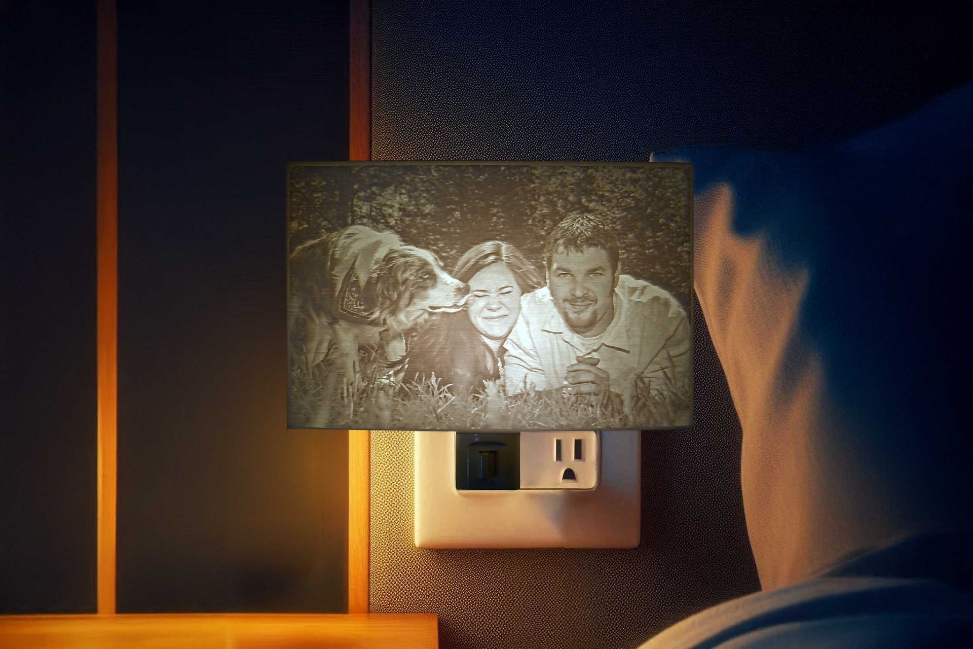 Nightstand photo frame above an electrical outlet, lit by warm bedside light.