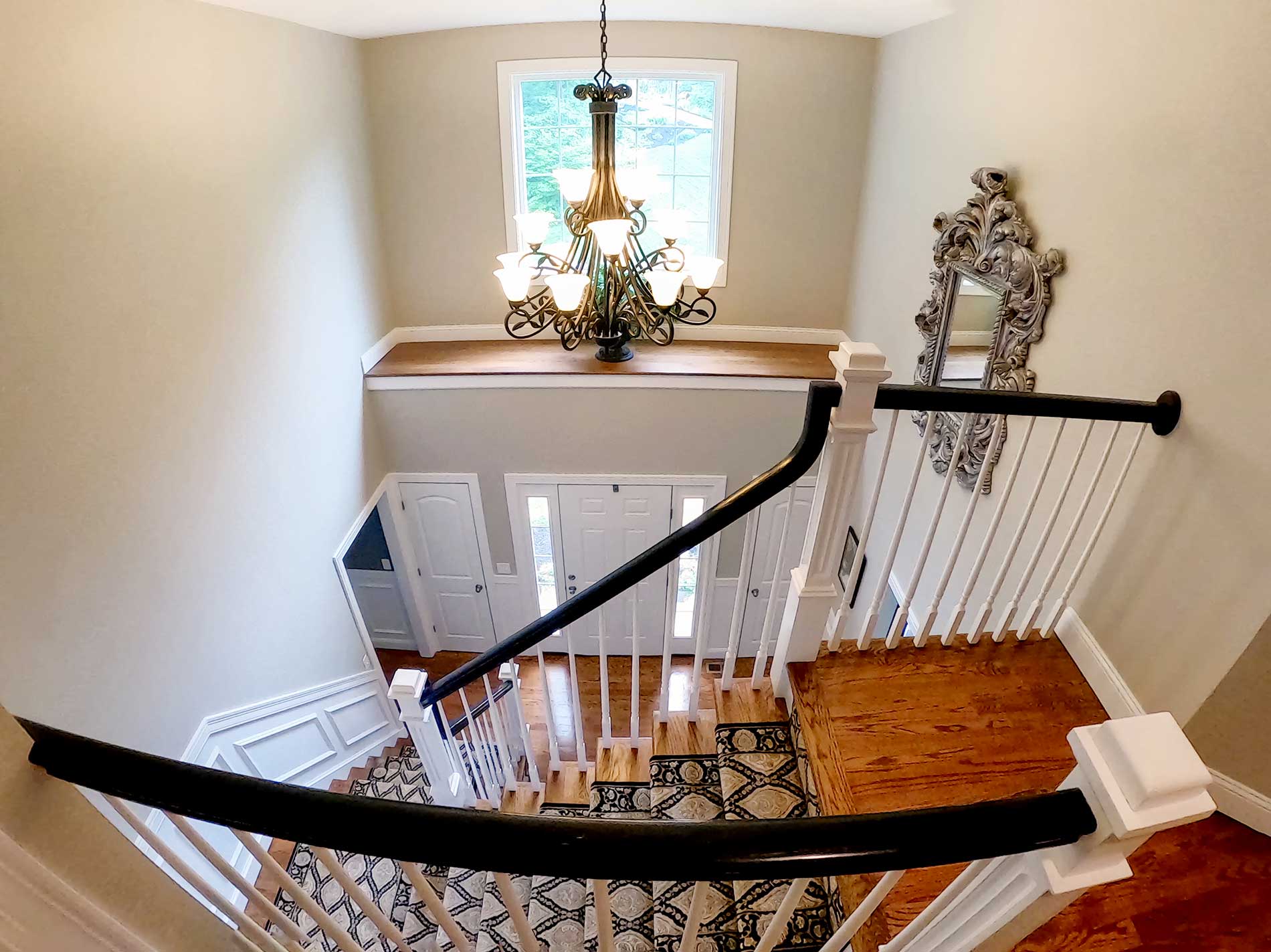 Staircase landing with chandelier, window, and curved black handrail over hardwood floor