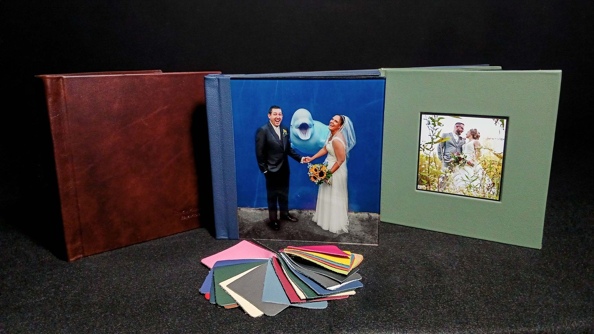 Three wedding album displays, one with a couple photo and scattered colorful paper samples on a dark surface