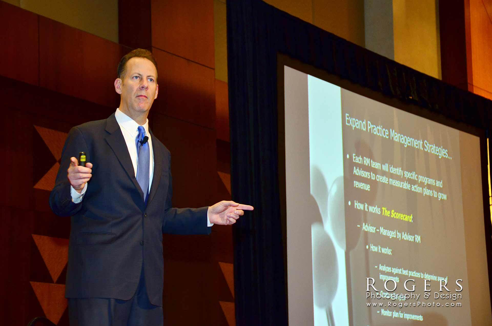 Presenter in a dark suit speaks beside a projected slide during a conference presentation.
