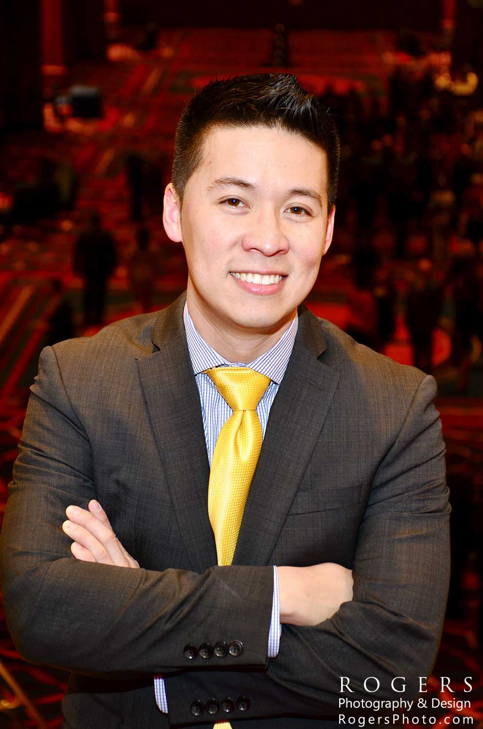 Smiling man in a gray suit and yellow tie with arms crossed in a dim red-lit setting