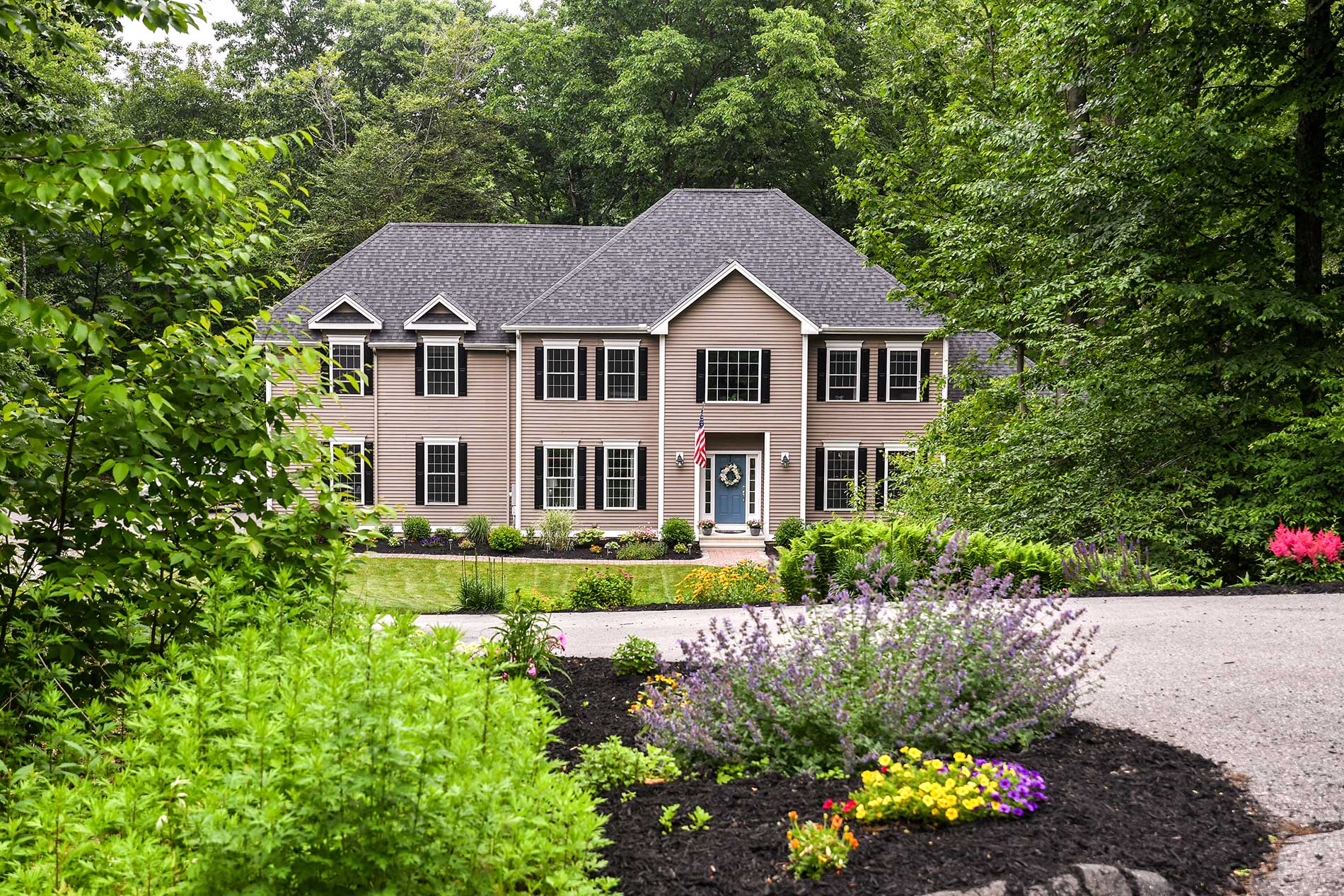 Two-story beige house with a front porch, surrounded by lush green trees and colorful garden flowers