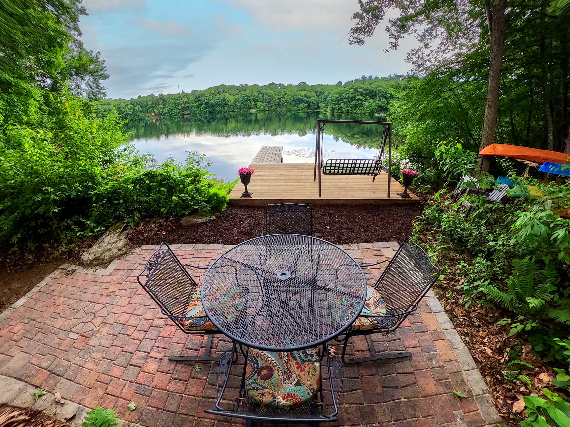 Brick patio overlooking a sandy lake beach with chairs, greenery, and a dock under trees