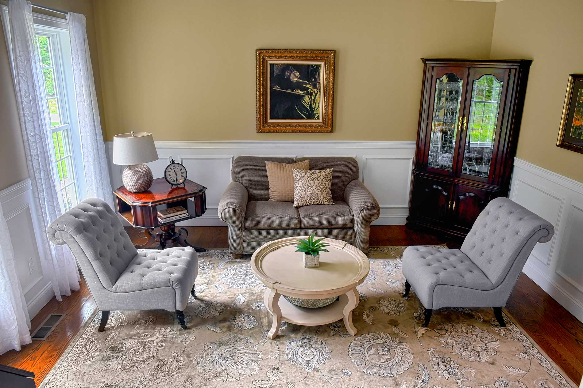 Bright sitting room with beige armchairs, sofa, coffee table, rug, and glass cabinet by a window.