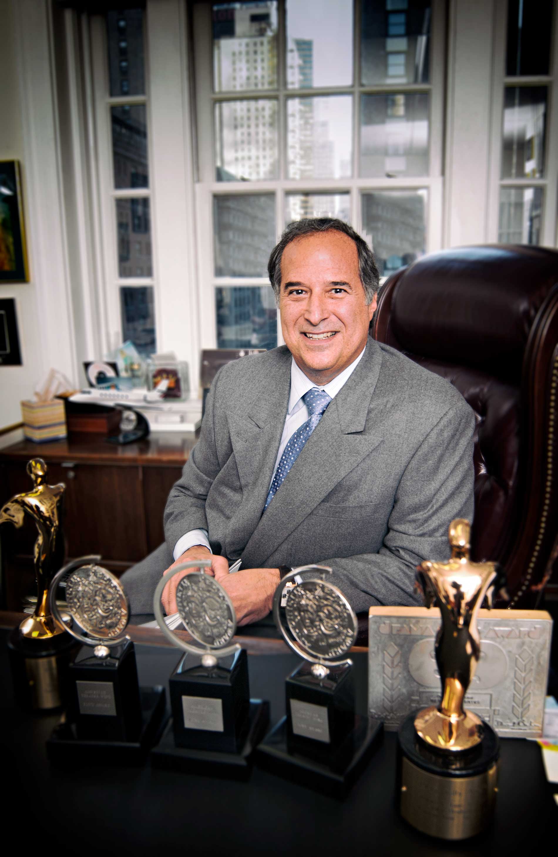 Stewart Lane sitting at a desk with several award trophies in an office, smiling at the camera.
