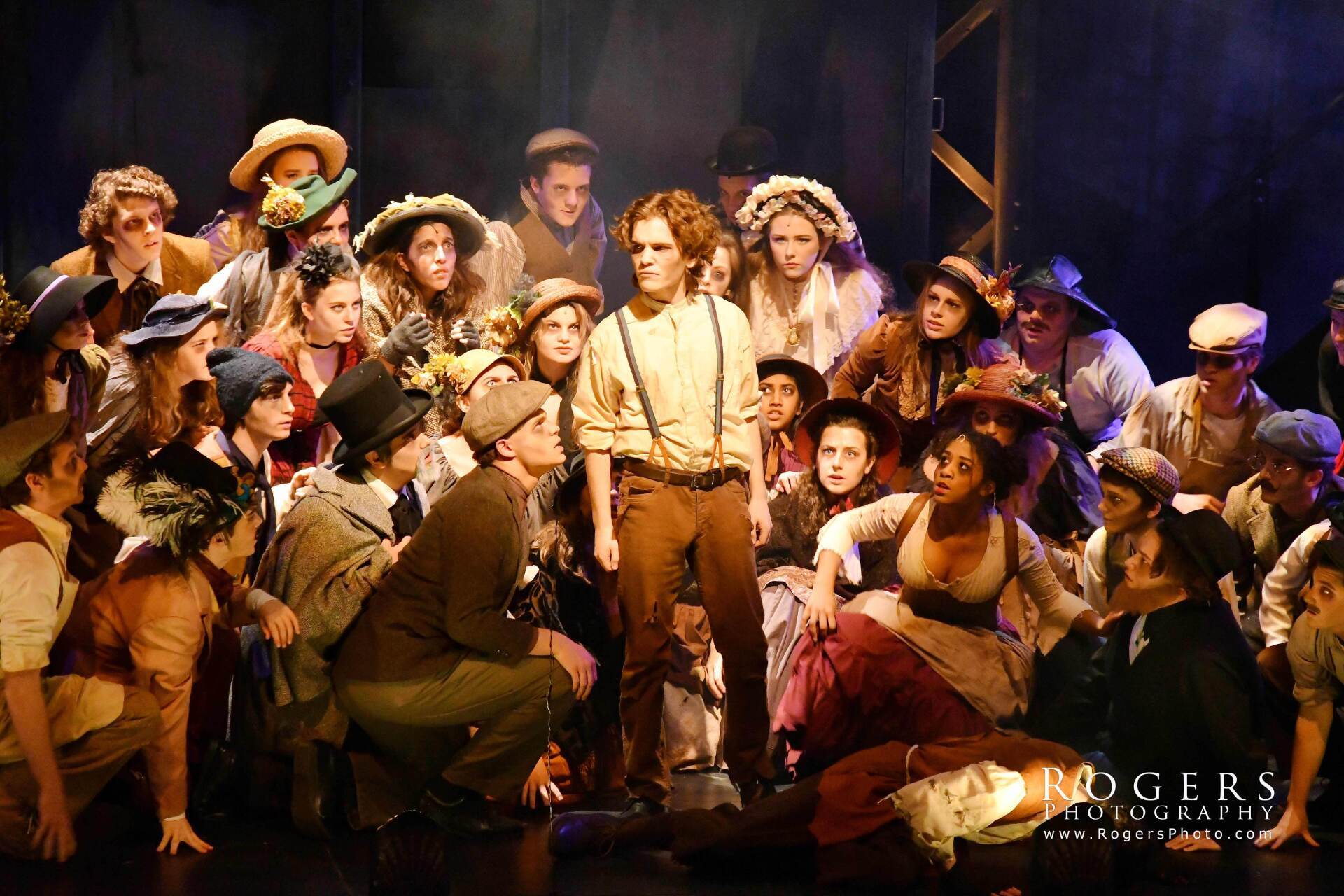 Theatrical cast in vintage costumes gathered on a dimly lit stage, singing and posing together in Sweeny Todd.