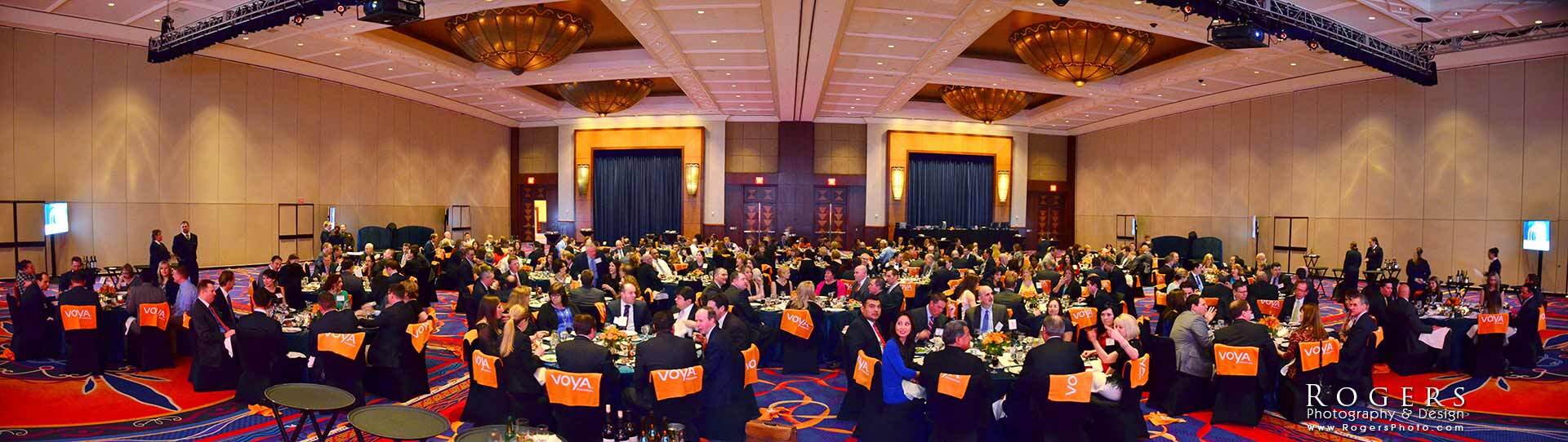 Large banquet hall filled with seated guests at round tables under warm chandeliers and ceiling drapes