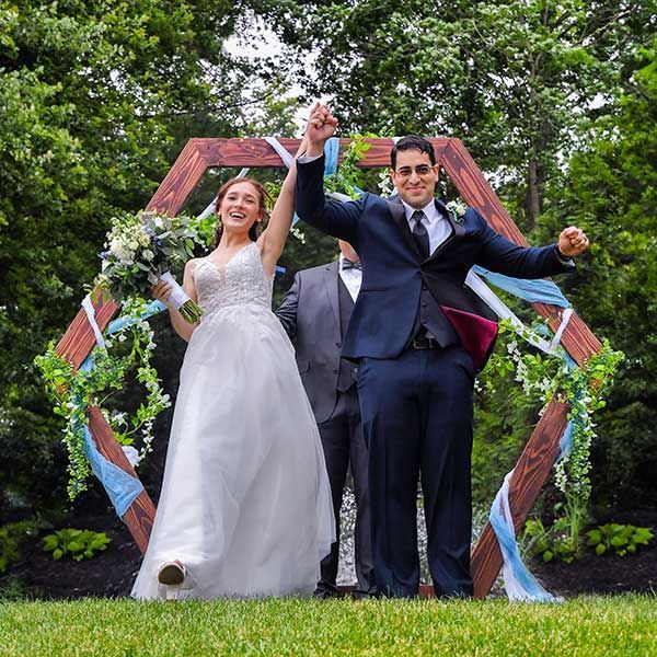 Newlyweds celebrate outdoors under a colorful hexagonal arch, hands raised and smiling.