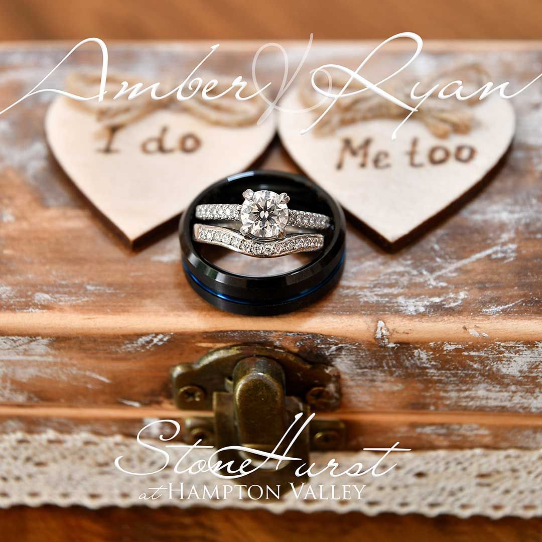 Wedding rings on a black box between heart notes reading “I do” and “Me too” on a rustic wooden table