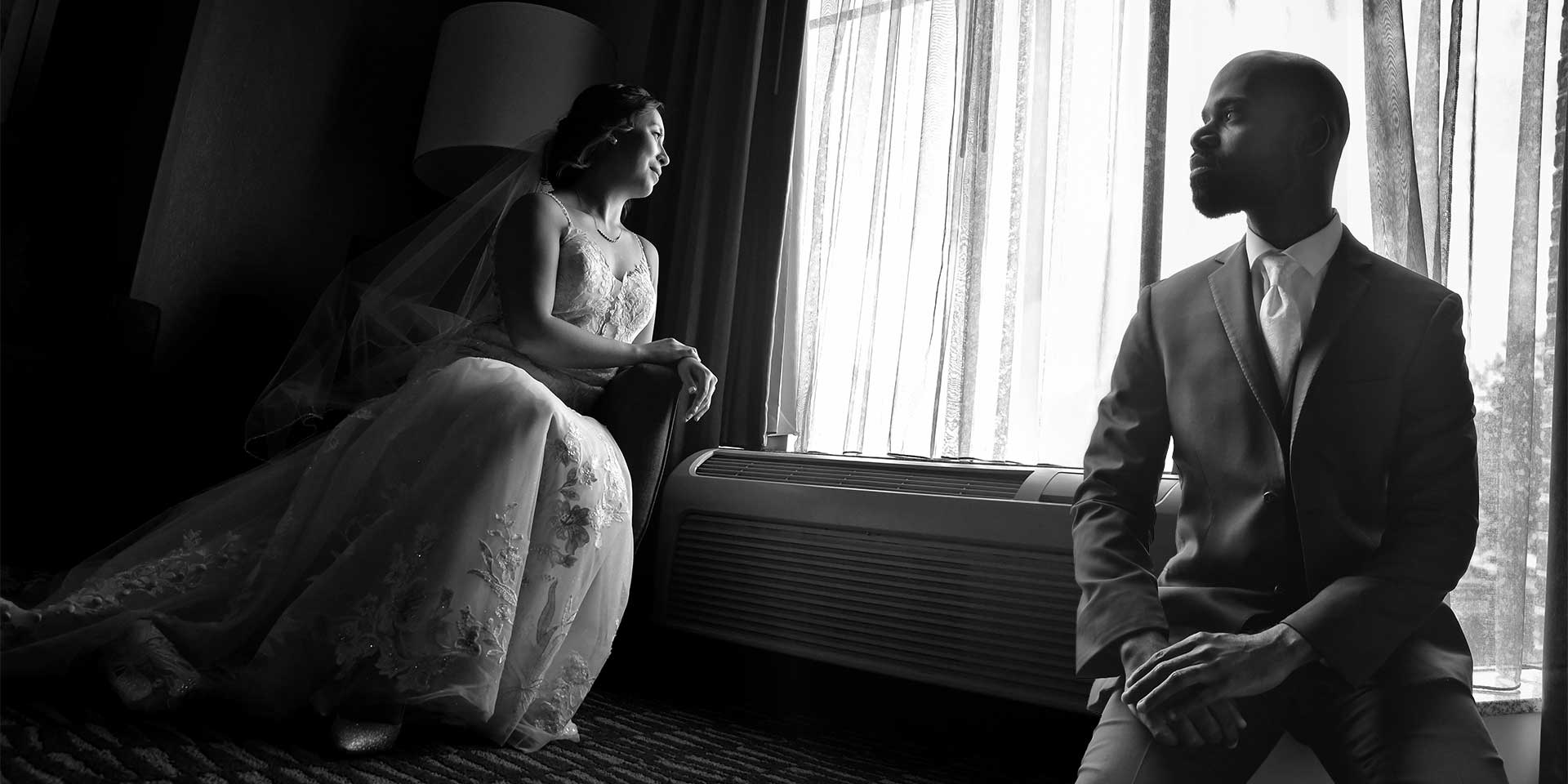 Bride and groom seated by a bright window, facing each other in a black-and-white scene