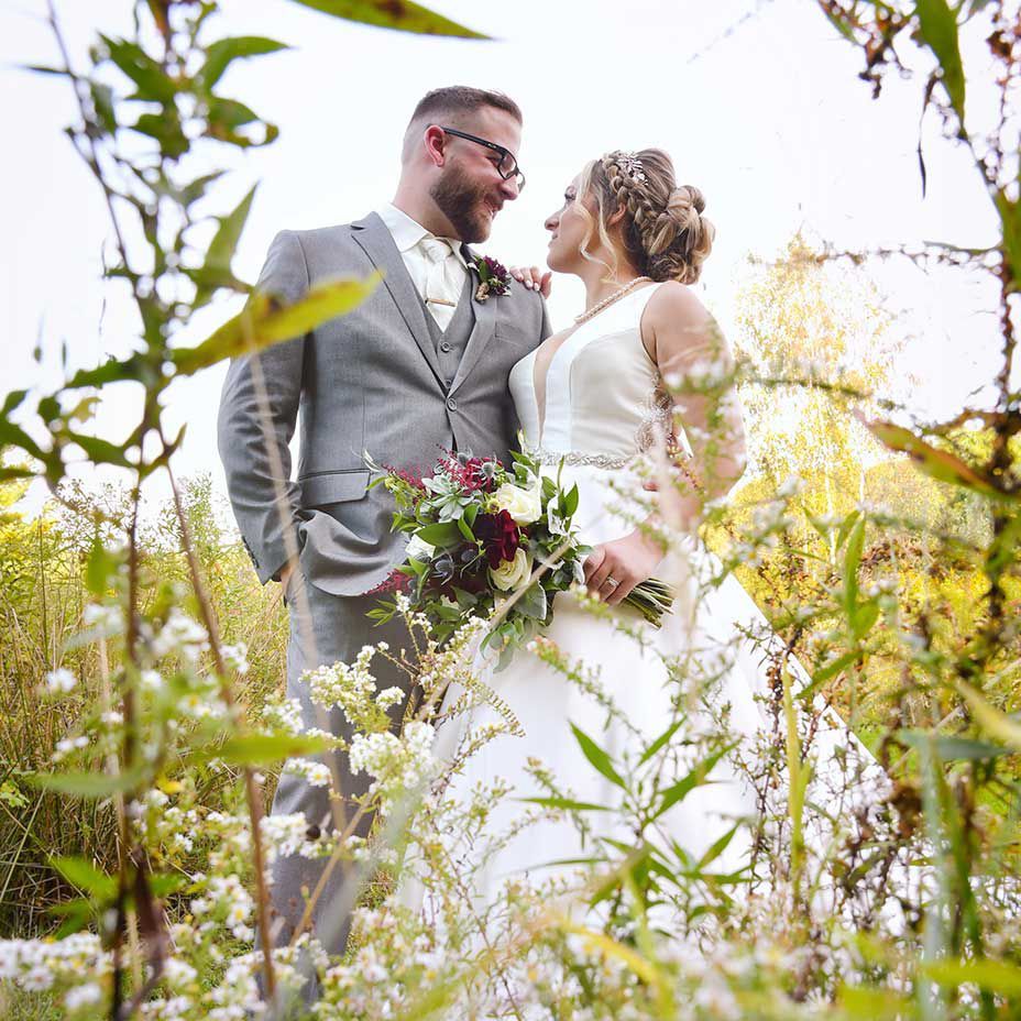 Bride and groom in a sunlit field, smiling and holding a bouquet of wildflowers.
