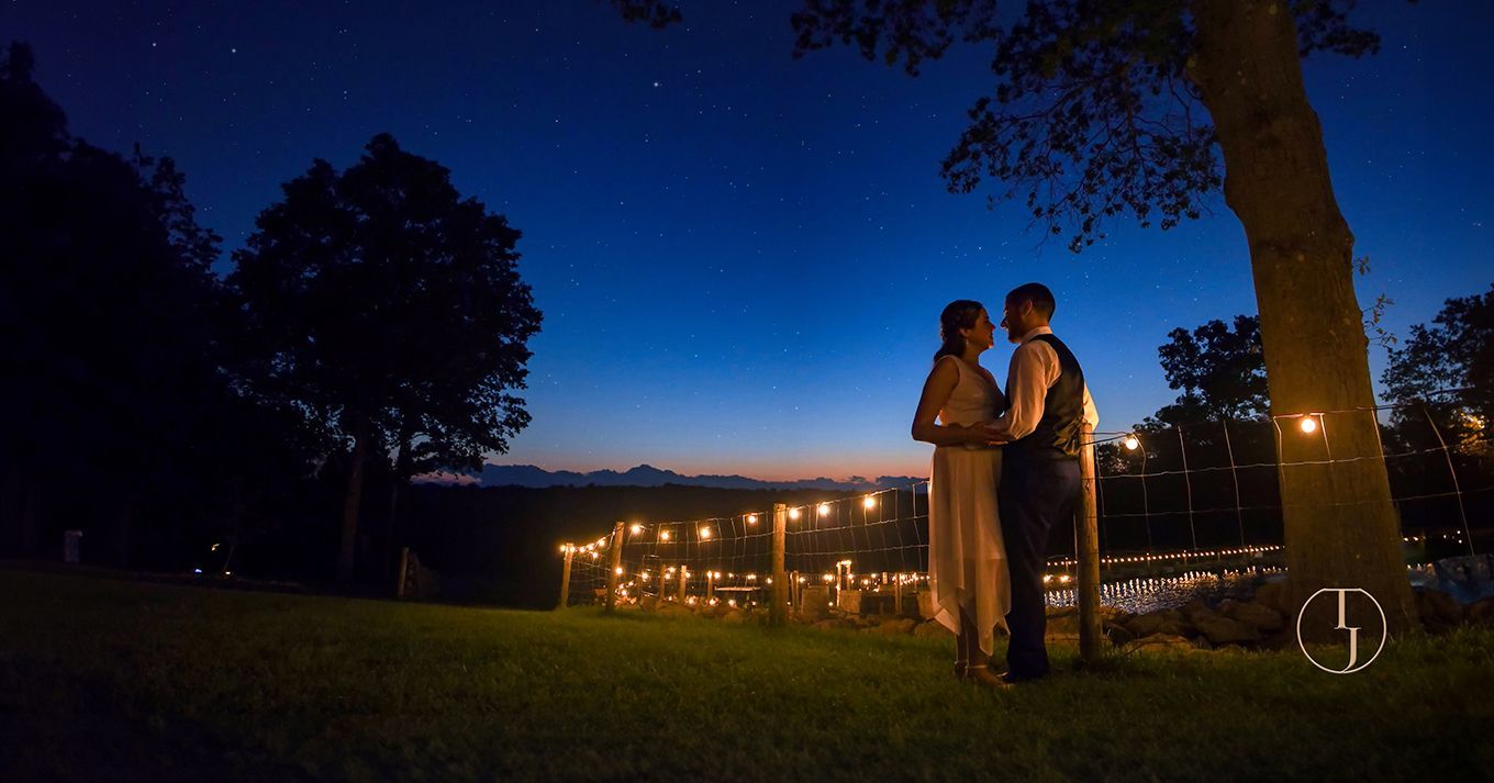 Couple standing on a lawn at dusk, with city lights and a warmly lit building in the background.