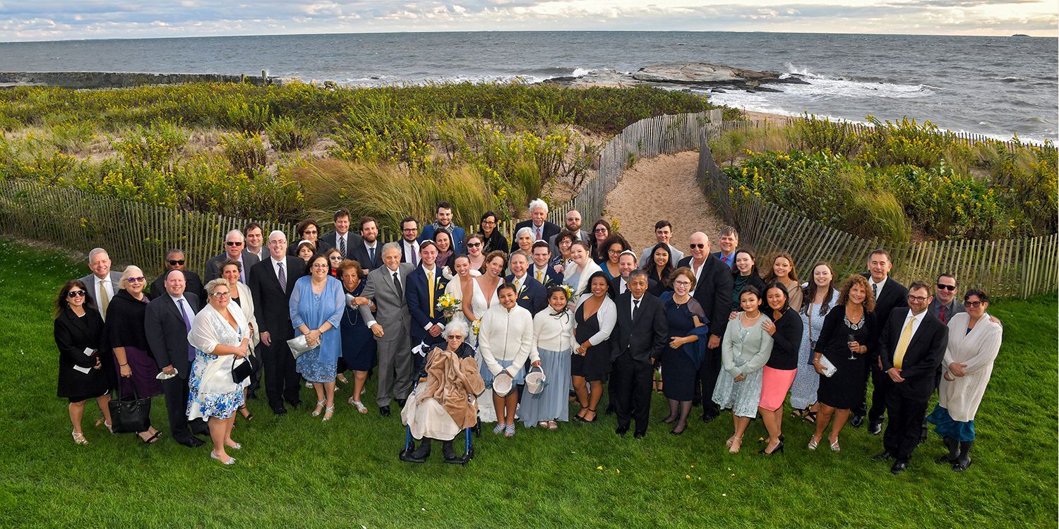 Large group posing outdoors on a grassy path with ocean and hills in the background, many waving and smiling