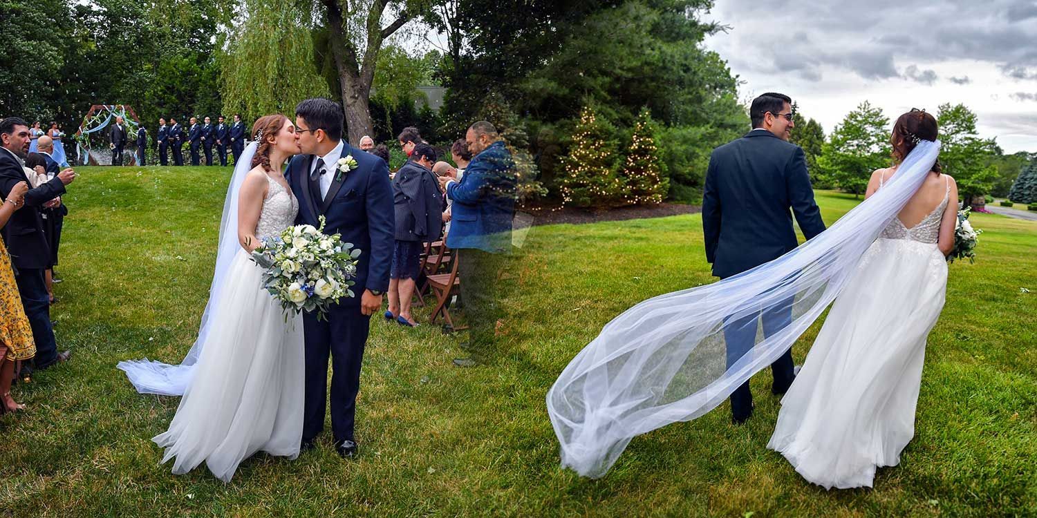Bride and groom standing on a lawn at a wedding, with her long veil blowing in the wind