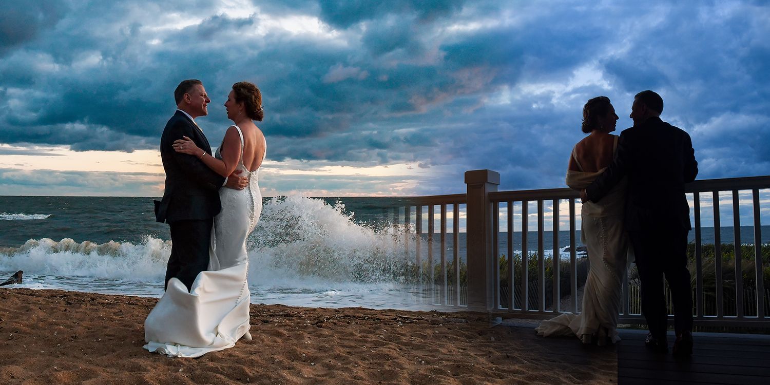 Bride and groom kissing by a waterfront railing at sunset, with waves crashing nearby