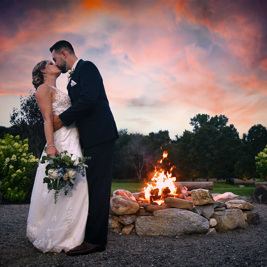 Wedding couple kissing beside a bonfire at dusk, with a pink-orange sky and garden trees in the background