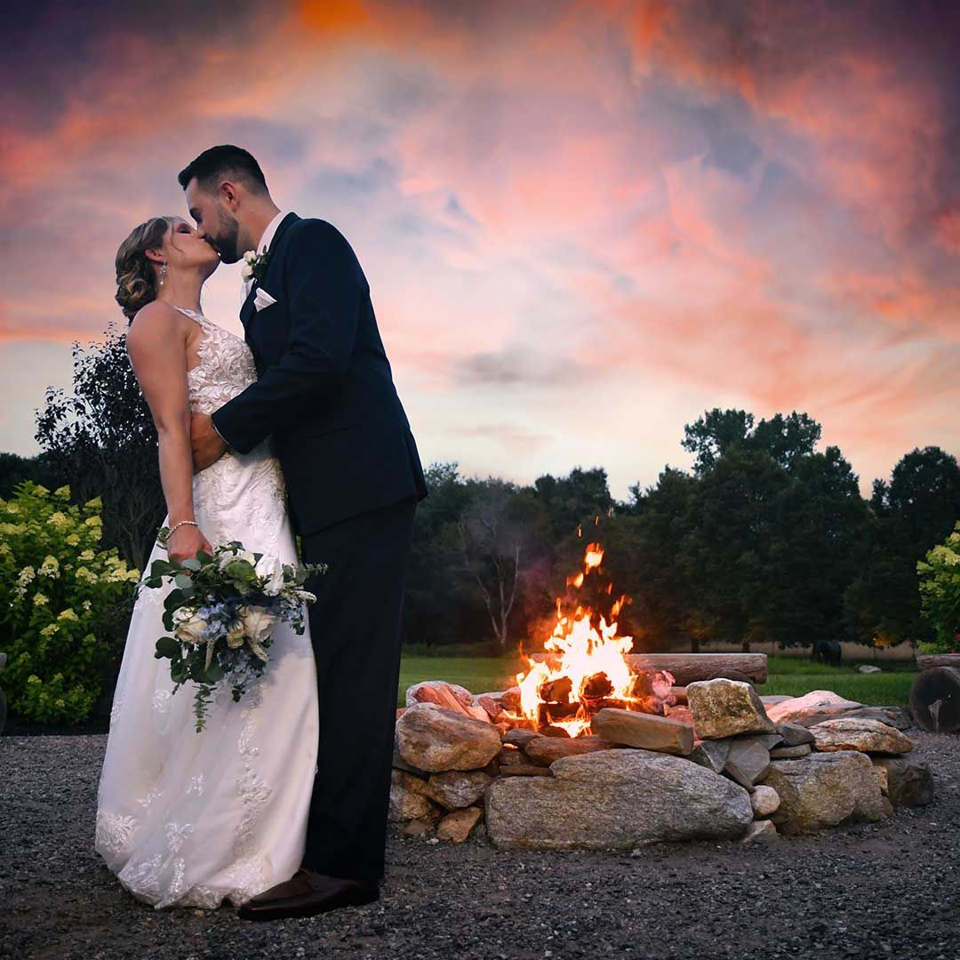 Bride and groom kiss beside a glowing campfire at sunset in an outdoor setting