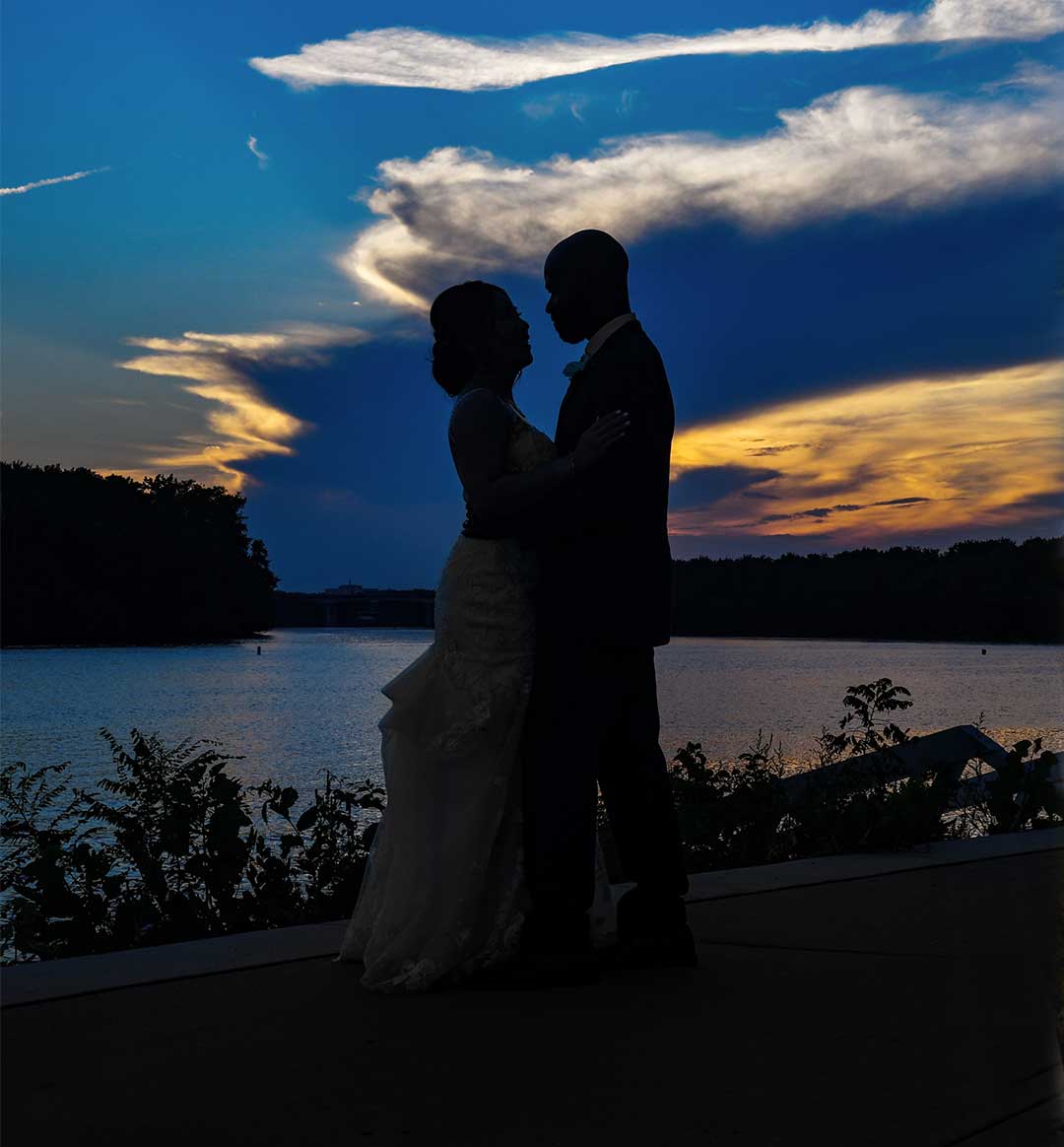 Silhouetted bride & groom embracing by a lake at sunset under dramatic blue-orange clouds