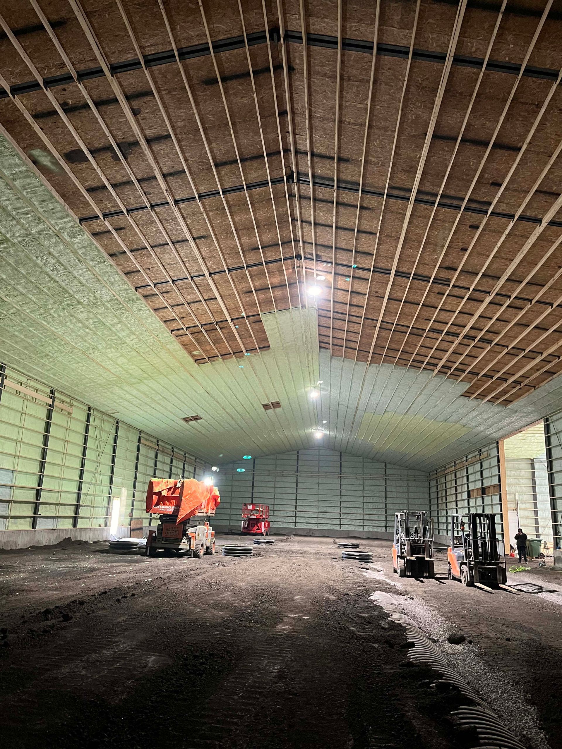 Large warehouse interior under construction, with exposed ceiling, dirt floor, and a small vehicle near the back.