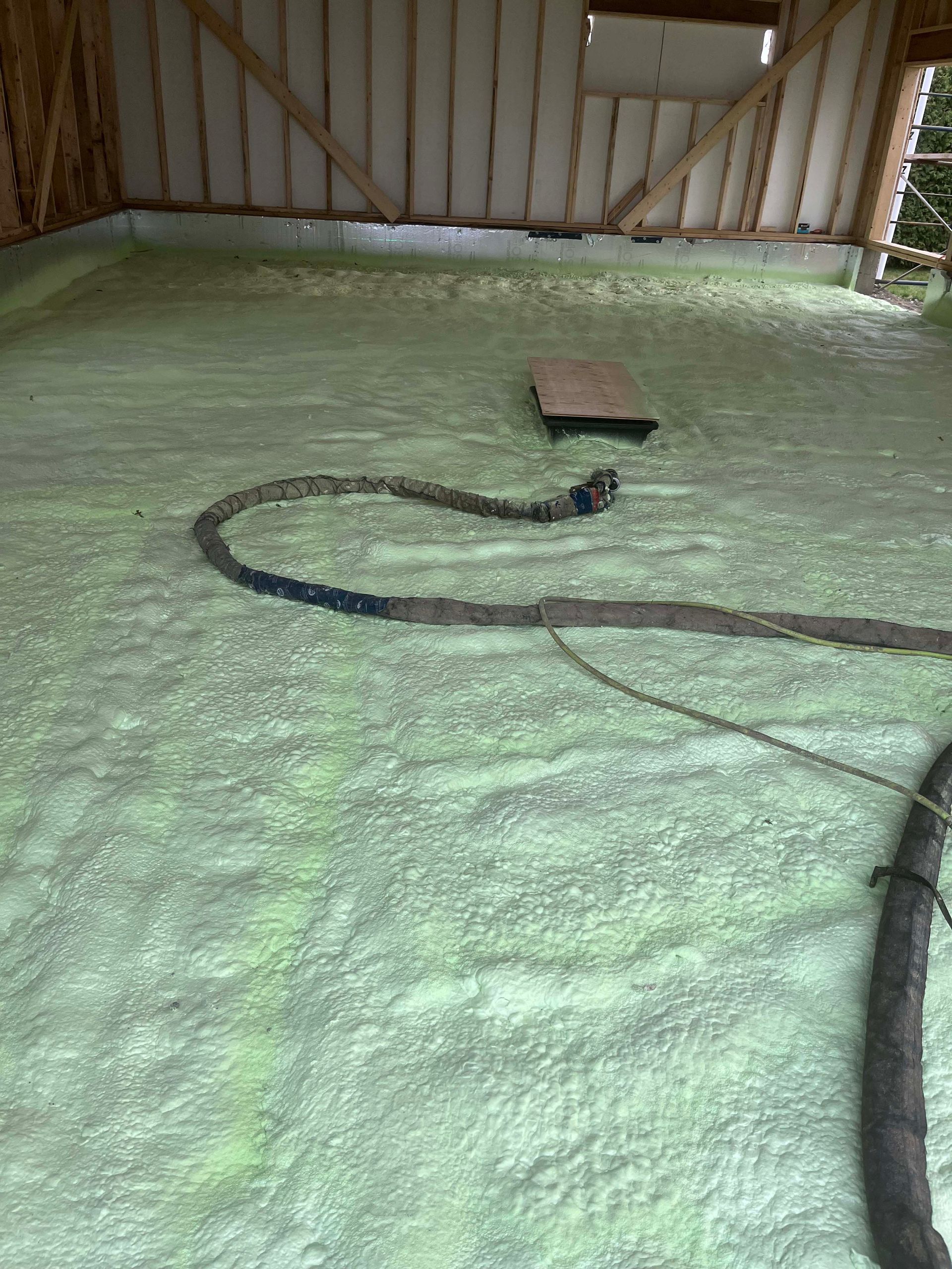 Insulated attic floor with green spray foam, exposed rafters, and a hose or cable lying across it