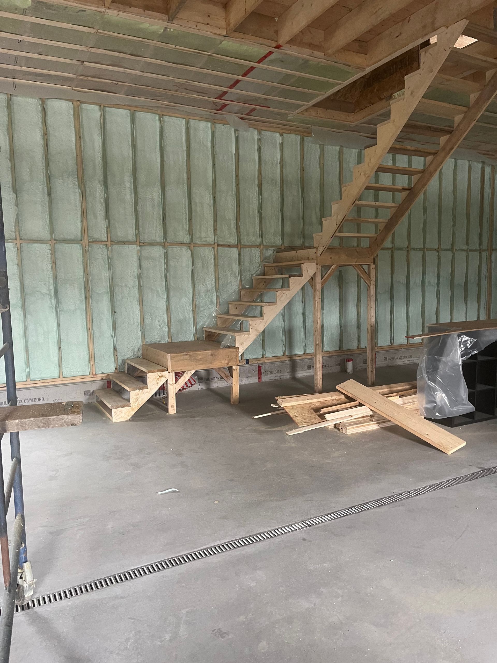 Interior of unfinished building with staircase, exposed insulation walls, and construction materials on concrete floor
