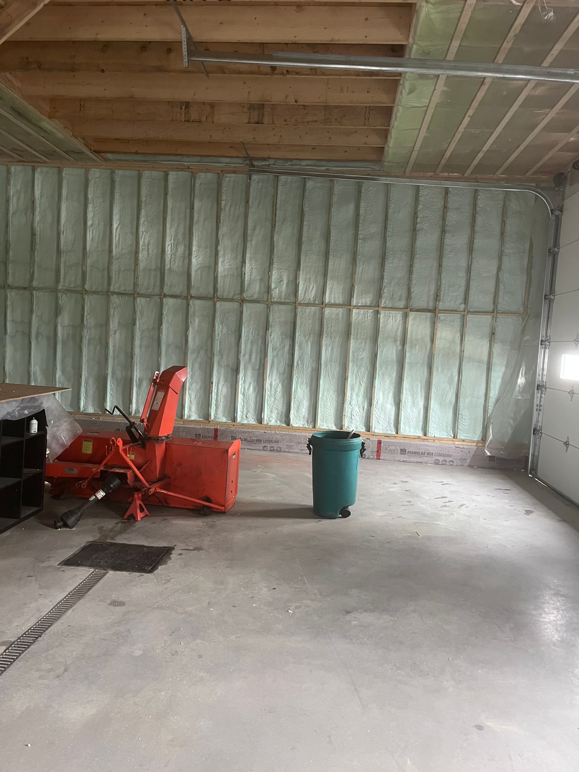 Insulated garage interior with concrete floor, exposed ceiling, orange snowblower, and green trash bin