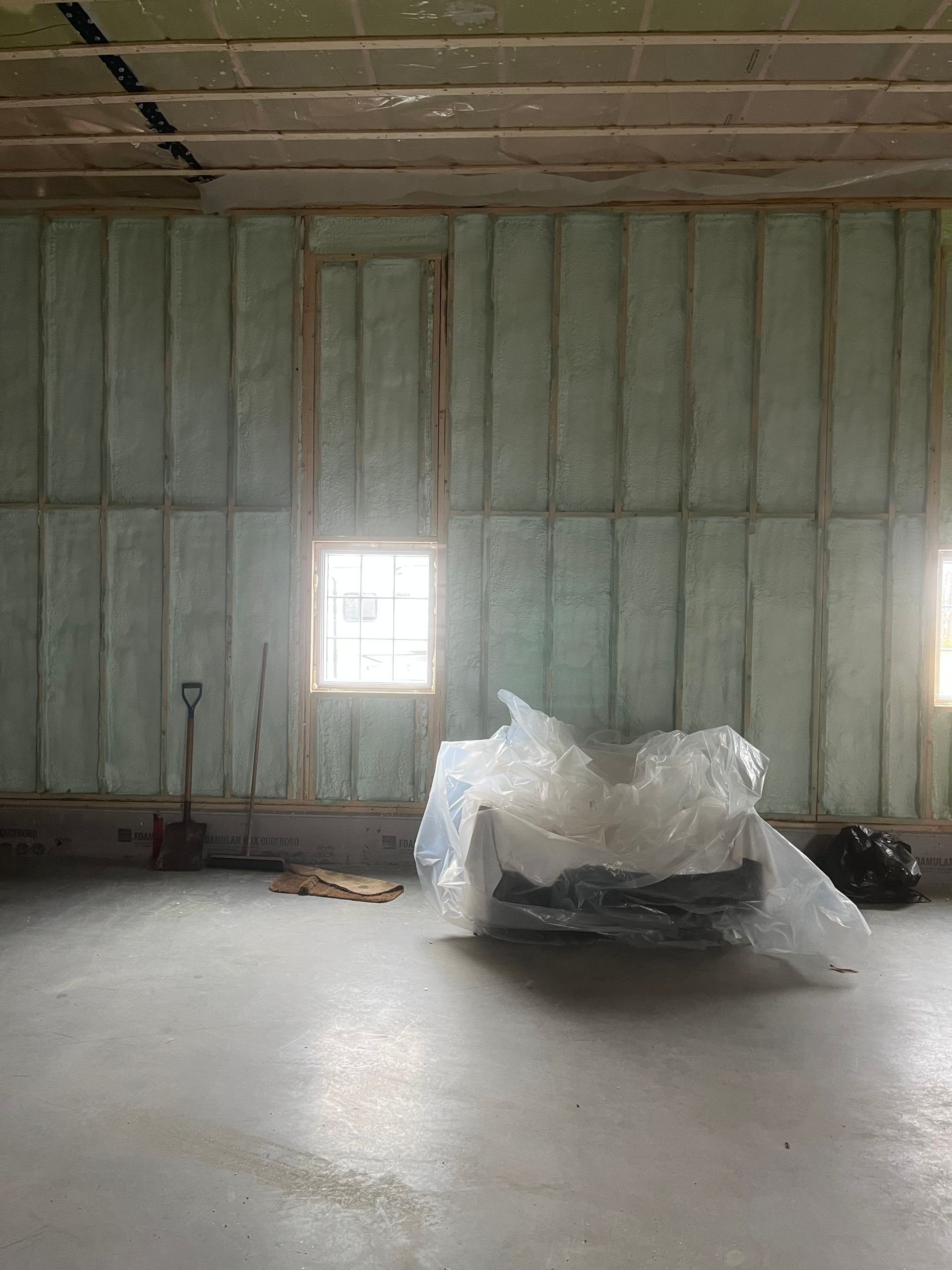 Empty unfinished room with insulated walls, ceiling beams, and a plastic-covered object on the floor near a window.