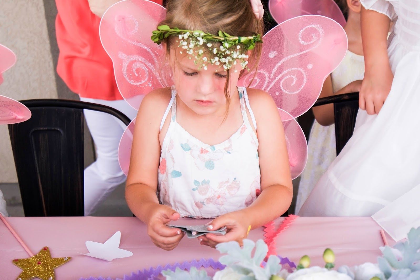 A little girl wearing fairy wings and a flower crown is sitting at a table.