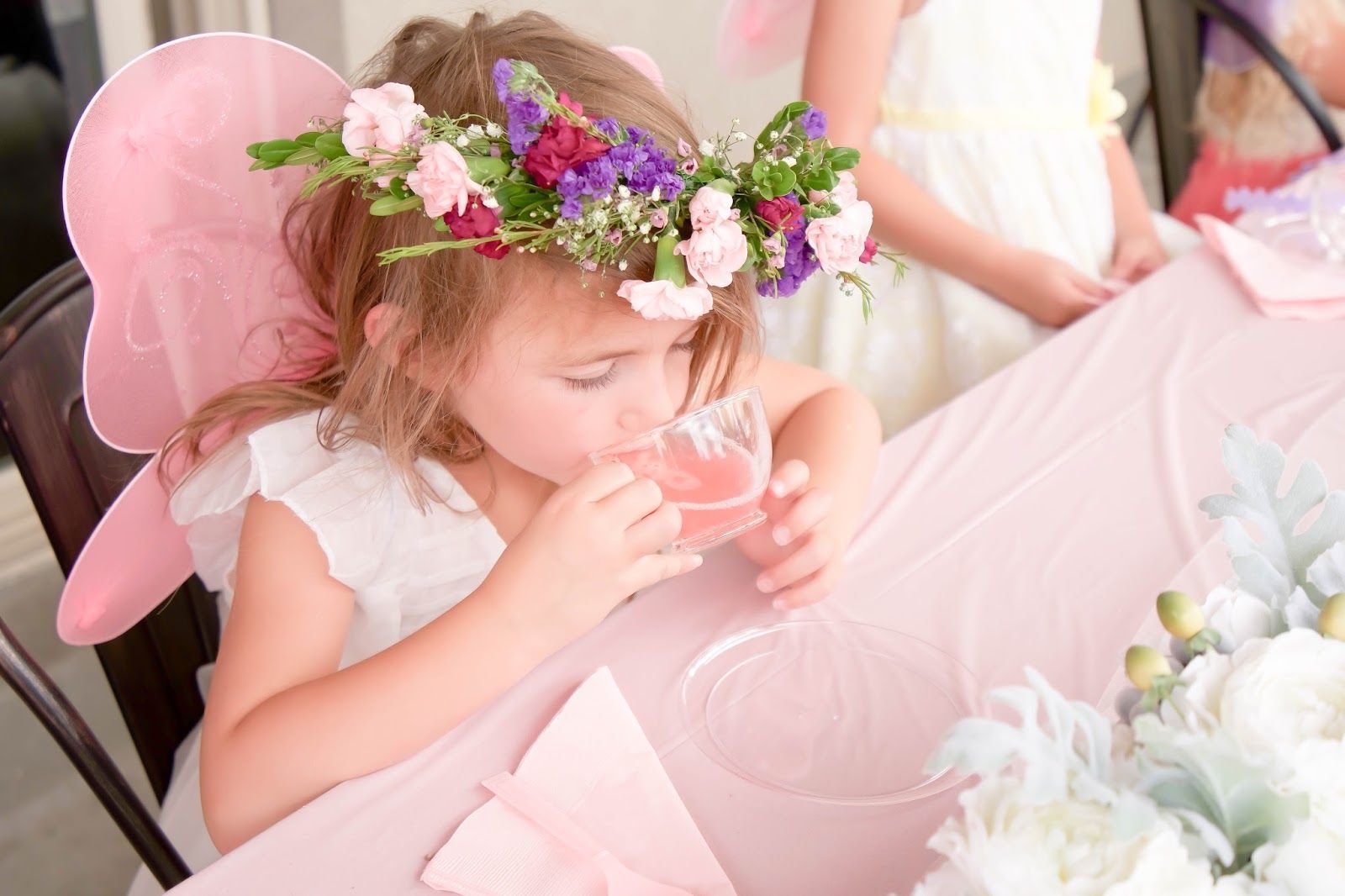 A little girl wearing a flower crown is drinking from a glass.
