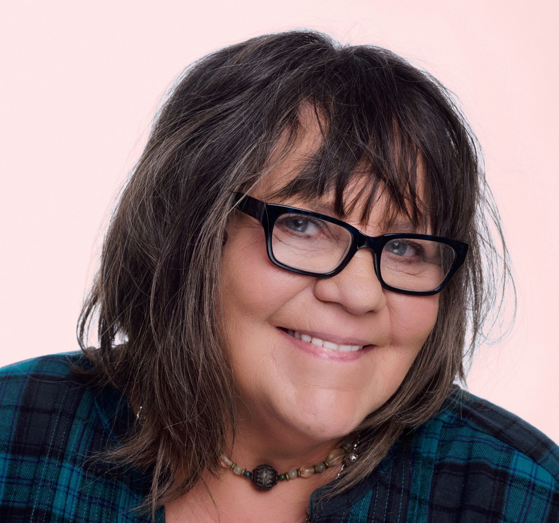 Woman with dark hair and glasses smiles; wearing a plaid shirt against a pink background.