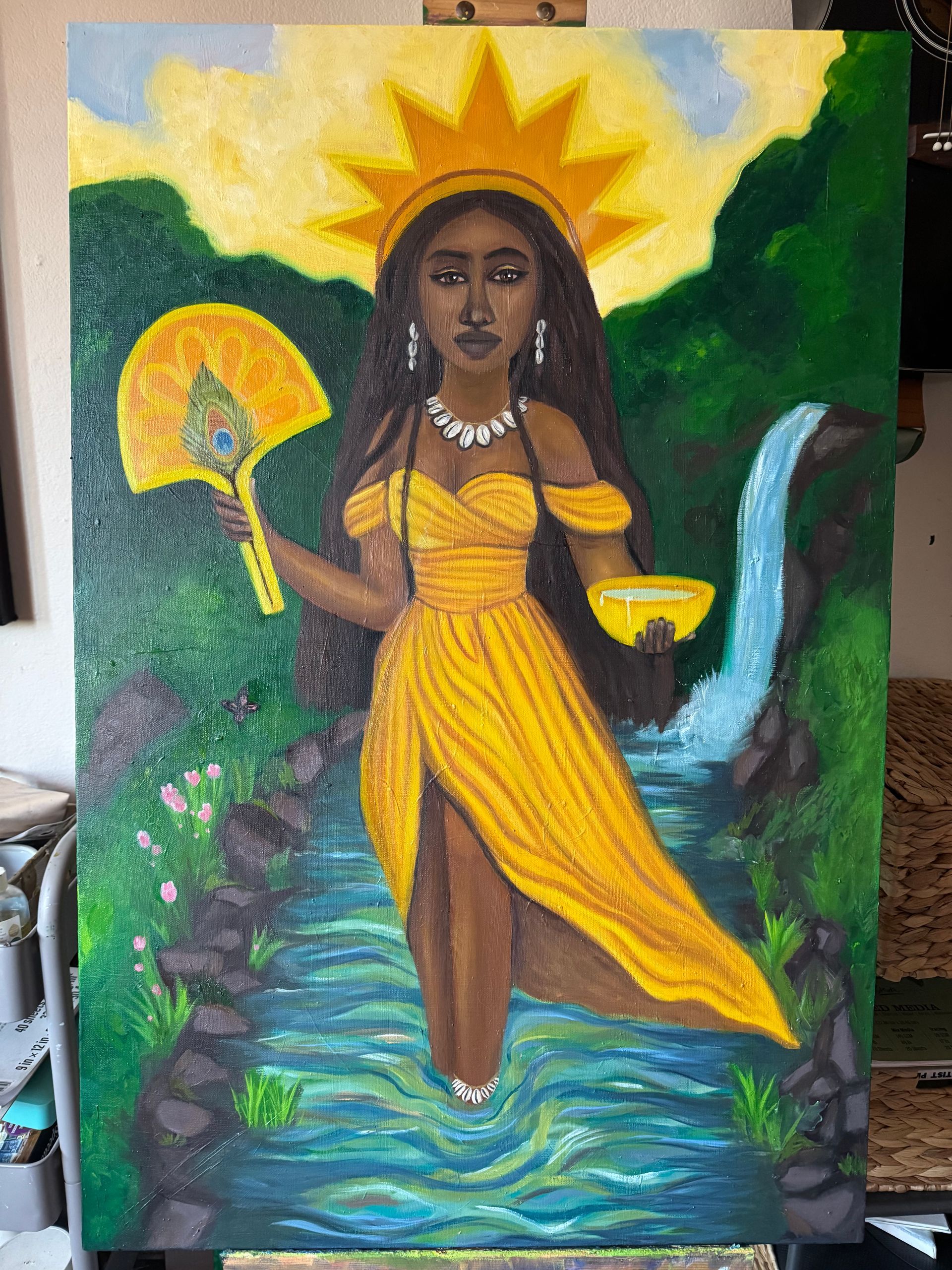 Painting of a woman in a yellow dress with a halo, holding a fan and bowl beside a waterfall and stream.