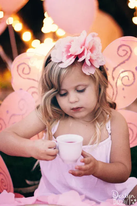 A little girl dressed as a fairy is pouring tea into a cup.